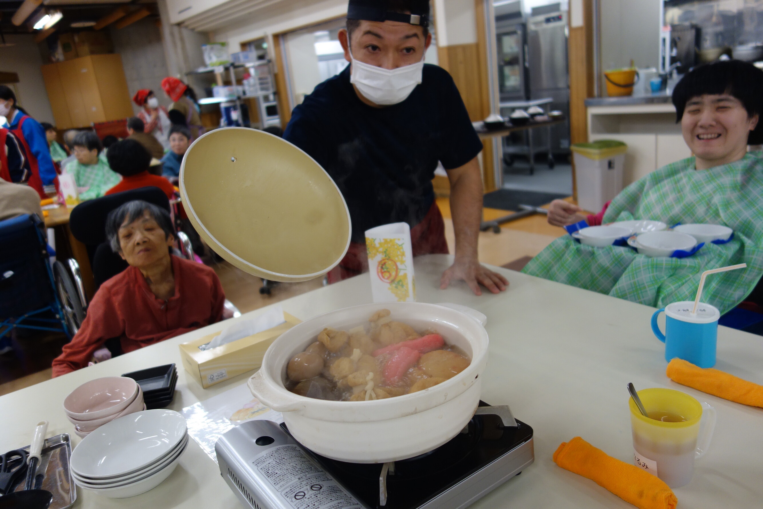 施設での鍋物会で熊田さんが利用者さんのテーブル上の鍋の蓋をパカっと開けている様子