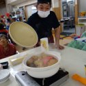 施設での鍋物会で熊田さんが利用者さんのテーブル上の鍋の蓋をパカっと開けている様子
